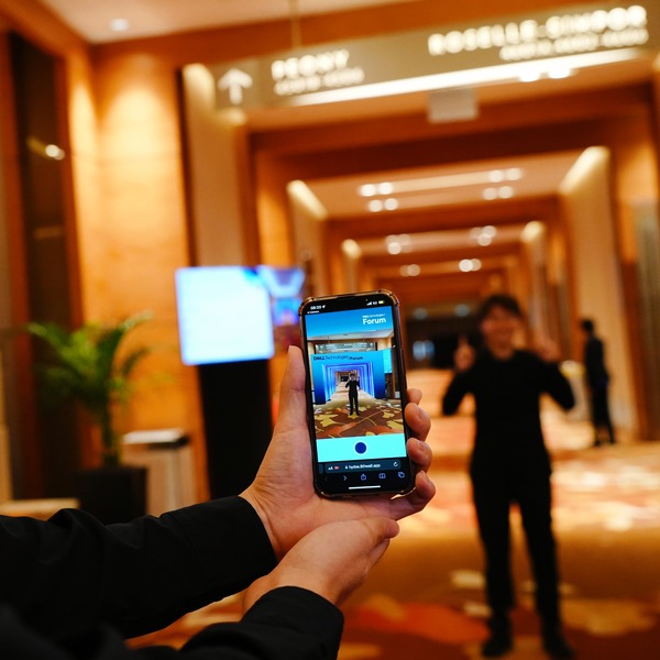 A pair of hands holding a smartphone that shows a man posing in front of a Dell Technologies Forum archway generated by AR technology.