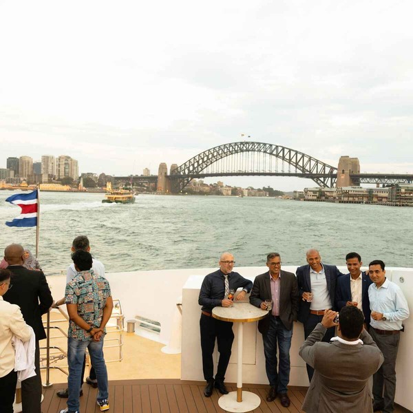 A lively group of Ericsson guests gathered on a boat's deck, sharing laughter and conversation against a backdrop of the Sydney Harbour Bridge.