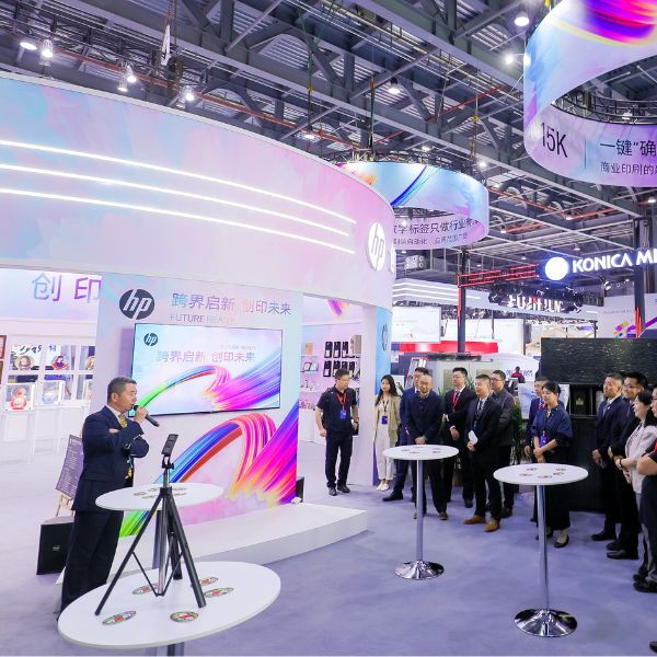 Print China 2023 event attendees gathering at tables outside the HP Indigo Print 15K Digital Press booth, viewing a speaker presentation.