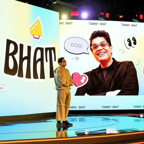 Tanmay Bhat speaking on stage, standing beside a large screen that displays a cheerful image of him.