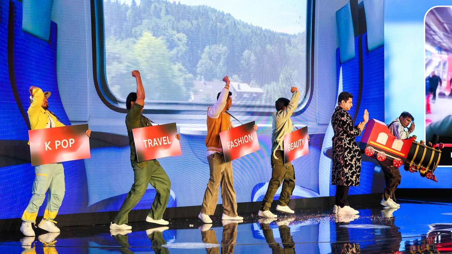 Performers on stage, joyfully lifting their arms, carrying red-coloured signs that say K-pop, Travel, Fashion and Beauty, creating an energetic atmosphere.